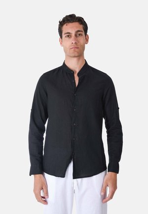 Uomo con camicia nera con collo mandarino abbottonata e pantaloni bianchi in piedi davanti a uno sfondo chiaro e uniforme.