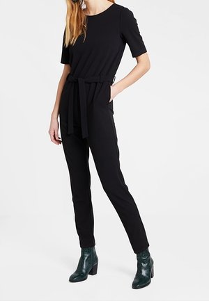 Žena v černém krátkém rukávu s opaskem, s úzkými kalhotami a tmavozelenými texturovanými anklet botami, stojící s jednou rukou v kapse.