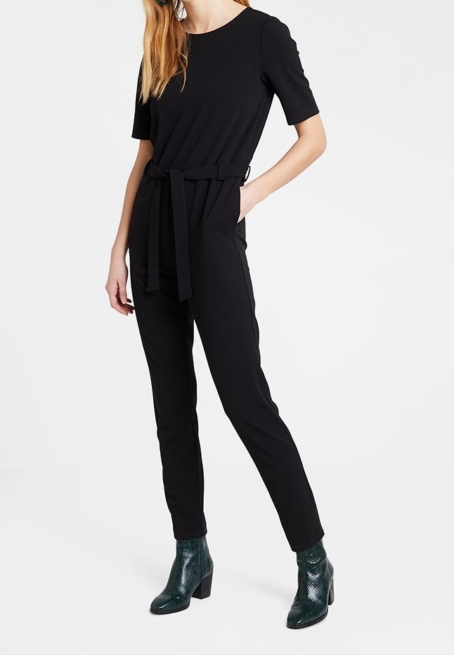 Femme portant une combinaison noire à manches courtes avec ceinture, avec des pantalons slim et des bottes à lacets noir texturé, se tenant avec une main dans la poche.