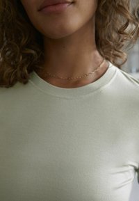 Nærbillede af en person iført en lysegrøn t-shirt med rund halsudskæring og en sart guldkæde om halsen, med krøllet hår der rammer nakken og skuldrene.