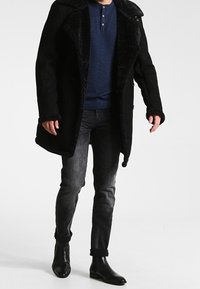Manteau en laine noire avec un col en fourrure, porté sur un t-shirt henley bleu foncé, assorti à un jean foncé et des bottines noires.