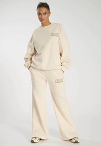 Conjunto de sudadera beige y pantalones a juego en tejido suave. La sudadera presenta el texto "PILATES SOUTH BEACH MIAMI". Diseño holgado y pierna ancha.