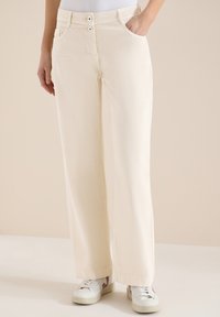 Femme portant un pantalon large crème à double fermeture boutonnée et des baskets blanches, une main dans la poche, sur un fond beige.