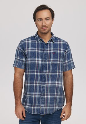 Mężczyzna ubrany w niebiesko-białą kratowaną koszulę na guziki z krótkim rękawem, stojący na jasnym, jednolitym tle, zwrócony przodem.