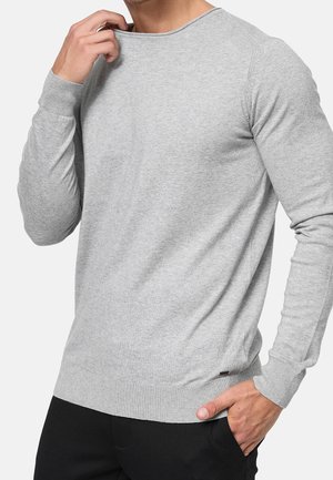 Uomo che sistema il colletto, indossando un maglione a maniche lunghe di colore grigio chiaro e pantaloni neri, con la mano in tasca su uno sfondo semplice.