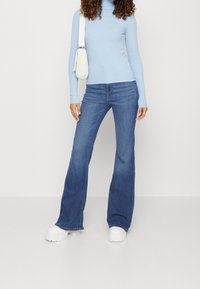 Lichtblauwe ribbel turtleneck trui gecombineerd met donkerblauwe uitlopende jeans. Model draagt witte chunky plateausneakers en een kleine witte handtas.