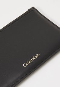 Zwarte lederen portefeuille met een gladde textuur, voorzien van een gouden bedrukte "Calvin Klein" logo en subtiele stiksels langs de randen.