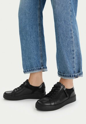 Füße in hochgekrempelten blauen Jeans und schwarzen flachen Leder-Sneakers mit seitlichem Reißverschluss und Schnürsenkeln auf hellem Hintergrund.