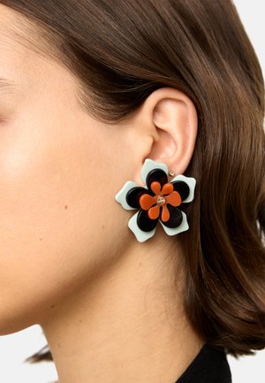 Lewe ucho kobiety z dużym kolczykiem w kształcie kwiatów o czarnych, białych i pomarańczowych płatkach na tle jasnej skóry i brązowych włosów.