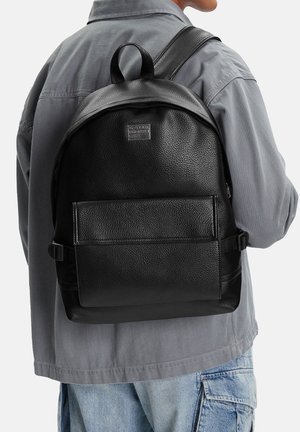 Person trägt einen schwarzen Rucksack aus strukturiertem Leder mit Vordertasche und grauer Jacke, kombiniert mit hellblauen Denim-Cargohosen.