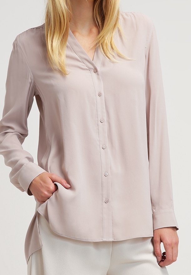 Femme portant un chemisier beige clair à manches longues, boutonné avec une poche latérale, associé à un pantalon blanc, debout devant un fond uni.