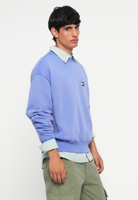 Sudadera de un suave color púrpura con puños acanalados, sobre una camisa a rayas en tonos azul claro y blanco. El modelo lleva pantalones cargo verde oliva.