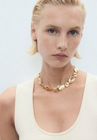 Gold necklace with irregular, oval-shaped links, varying in size and texture, worn over a light-colored top. Smooth and shiny finish.