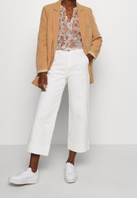 Blazer beige, blusa floral con patrones rojos y verdes, pantalones blancos anchos y cortos, y zapatillas blancas con suelas texturizadas.