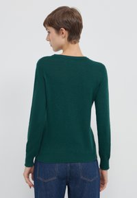 Frau mit kurzen braunen Haaren, die einen dunkelgrünen, langärmeligen Pullover und blaue Jeans trägt, von hinten gegen einen einfarbigen grauen Hintergrund blickend.