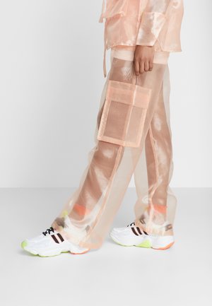 Person iført gennemsigtige ferskenfarvede bukser med en stor lomme, kombineret med hvide sneakers med sorte og orange striber og neongrønne såler.