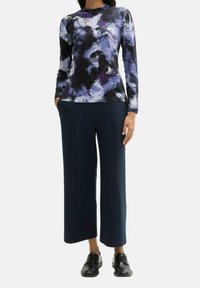 Dlouhý rukáv vrchního dílu s vícebarevným abstraktním potiskem v odstínech fialové a černé; zkombinováno s tmavě modrými širokými kalhotami; model má na sobě černé boty.