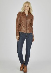 Braune Lederjacke mit einem Reißverschluss vorne, zwei Brusttaschen und enganliegenden Bündchen. Kombiniert mit einem weißen Oberteil, dunklen Jeans und braunen Stiefeln.