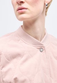 Κοντινό πλάνο ενός ατόμου που φοράει ένα απαλό ροζ καπιτονέ μπουφάν με κουμπί σναπ και ασ ημένιο κρίκο στο αυτί, με ορατά τα χείλη και το σαγόνι.