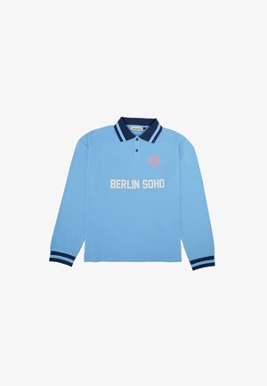 Polo a maniche lunghe blu chiaro con colletto e polsini blu scuro, presenta la scritta bianca "BERLIN SOHO" e un logo circolare sul lato sinistro del petto.