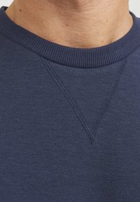 Marineblauwe sweatshirt, zachte stof, ribbelachtige halslijn, hoekige schoudernaad, subtiele stikseldetails, gladde textuur en klassiek ontwerp.