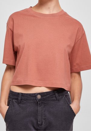 Persoon die een korte, terracotta T-shirt met korte mouwen draagt, lichtjes ingestopt in een hoog getailleerde donkergrijze spijkerbroek, met handen in de zakken.