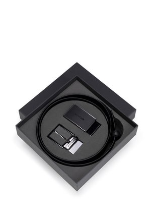 Czarny skórzany pasek zapakowany w kwadratowe czarne pudełko, z teksturowaną czarną sprzączką i srebrnym metalowym akcentem. Zestaw zawiera dodatkową sprzączkę.