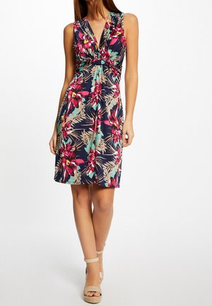 Femme portant une robe sans manches bleu marine à imprimé floral rose et bleu sarcelle, longueur genou, associée à des sandales compensées beiges à bride à la cheville.