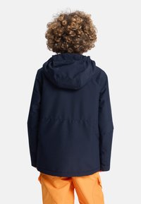 Granatowa wodoodporna kurtka z kapturem, o gładkiej fakturze i elastycznych mankietach. Noszona z jaskrawopomarańczowymi spodniami.