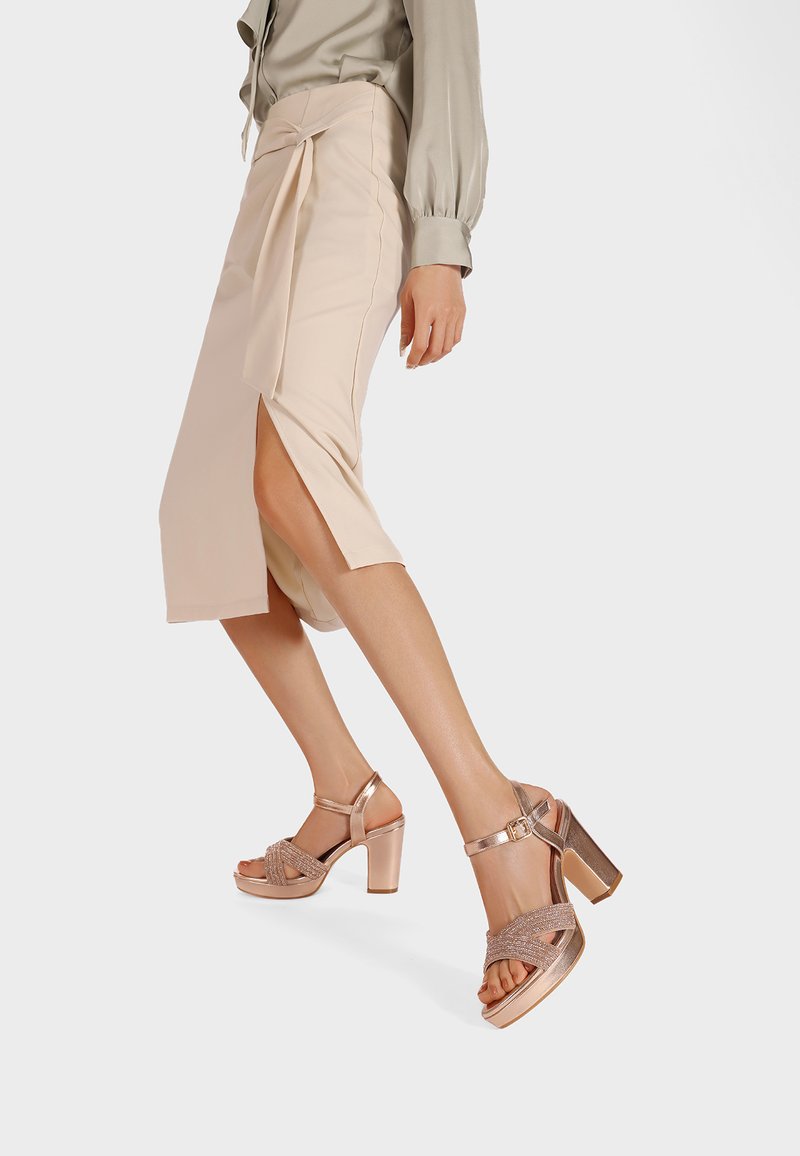 Falda midi beige con abertura lateral y detalle de lazo, combinada con sandalias de tacón bloque metalizadas en rosa dorado con correas trenzadas.