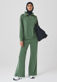 Green, zippered sweatshirt with a collar paired with matching wide-leg pants. Model holding a black quilted bag. White sneakers.
