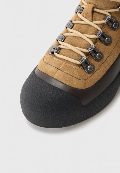 Zapato de escalada de ante marrón con una punta de goma negra texturizada, cordones beige, ojales metálicos y acentos cosidos en la parte superior.