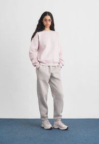 Φώλια ροζ φούτερ με ριγωτούς καρπούς, συνδυασμένο με γκρι φόρμες. Το μοντέλο φοράει ανοιχτόχρωμα αθλητικά παπούτσια και μωβ γυαλιά ηλίου. Απλός σχεδιασμός.