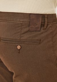 Close-up of brown textured pants with back button pocket and leather brand label, worn with beige waffle-knit top.