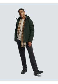 Green hooded puffer jacket, light beige sweater, plaid scarf in brown tones, black pants, and black shoes. Standing pose on a light background.