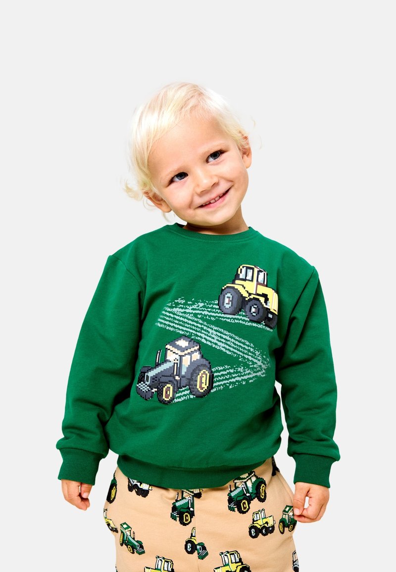 Enfant blond souriant et penchant la tête, portant un sweat-shirt vert et un pantalon beige avec des motifs colorés de tracteurs sur les deux vêtements.