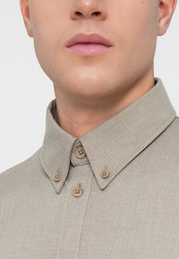 Μπεζ πουκάμισο σε σχέδιο ψαροκόκαλο με κατιόν κολάρο, που διαθέτει καφέ κουμπιά. Το ύφασμα έχει μια διακριτική υφή, τονίζοντας το υoveμένο σχέδιο.