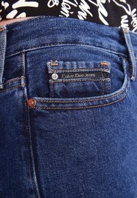 Tummansiniset farkut oransseilla ompeleilla. Ominaisuuksiin kuuluu pieni takatasku, jossa on merkintä "Calvin Klein Jeans" sekä metalliset niitit koristeena.
