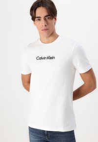 Camiseta de algodón blanca con cuello redondo y mangas cortas, que presenta el logotipo "Calvin Klein" en impresión negra en el pecho.