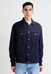 Mörk denimjacka med knappstängning, två bröstfickor och detaljer i gul sömnad. Har en klassisk skjortkrage och långa ärmar.
