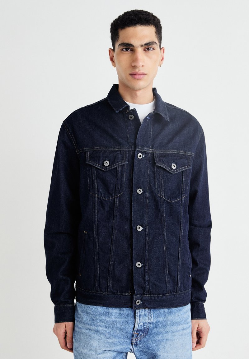 Mörk denimjacka med knappstängning, två bröstfickor och detaljer i gul sömnad. Har en klassisk skjortkrage och långa ärmar.