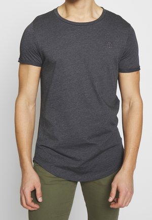 Mann trägt ein eng anliegendes, dunkelgraues T-Shirt mit kurzen Ärmeln und olivgrüne Hosen, steht mit locker an den Seiten hängenden Armen.