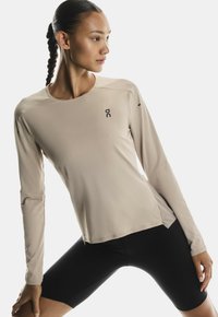 Beige Langarm-Athletikoberteil mit rundem Halsausschnitt, strukturiertem Stoff und kleinem schwarzem Logo auf der Brust. Kombiniert mit schwarzen Shorts.