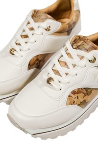 Sneakers bianchi con accenti in colore beige a motivo mappa, lacci bianchi e occhielli decorativi in oro con charm per i lacci, mostrati in primo piano.