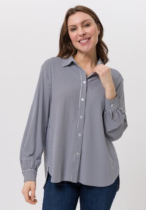 Lachende vrouw die een zwart-wit verticale gestreepte blouse met knopen en een blauwe spijkerbroek draagt tegen een effen lichte achtergrond.