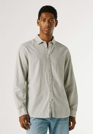 Giovane uomo che indossa una camicia a maniche lunghe grigio chiaro con motivo e jeans blu, in piedi davanti a uno sfondo uniforme.