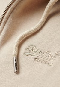 Tejido de sudadera color beige claro con una superficie texturizada, que presenta un zipper metálico plateado y un cordón crema. Logo bordado de "Superdry".