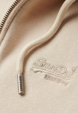 Lys beige hoodie-stof med en tekstureret overflade, der har en sølvmetal lynlås og en cremefarvet snor. Broderet "Superdry" logo.