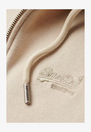Lys beige hoodie-stof med en tekstureret overflade, der har en sølvmetal lynlås og en cremefarvet snor. Broderet "Superdry" logo.