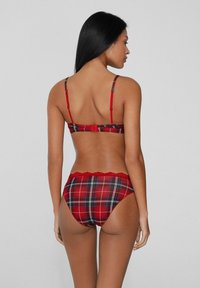 Mujer de espaldas, de pie, con un conjunto de sujetador y ropa interior de cuadros rojos y detalles de encaje en la cinturilla de la ropa interior, sobre un fondo liso.